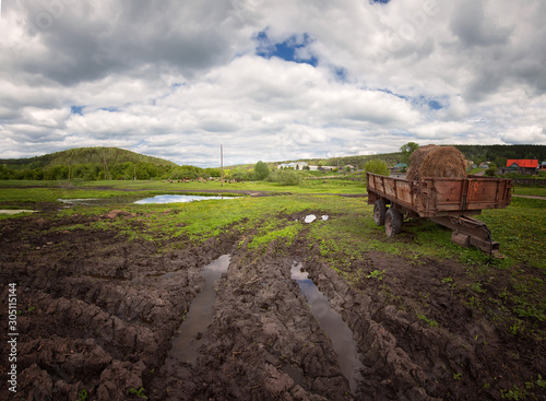 hay season, summer landscape old iron trailer with a roll of hay on a dirty village road, white clouds in the blue sky,