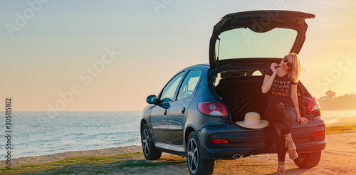 Young woman sits in a car trunk by the sea on the beach. Summer, sunset, road traveling concept. Copy space.