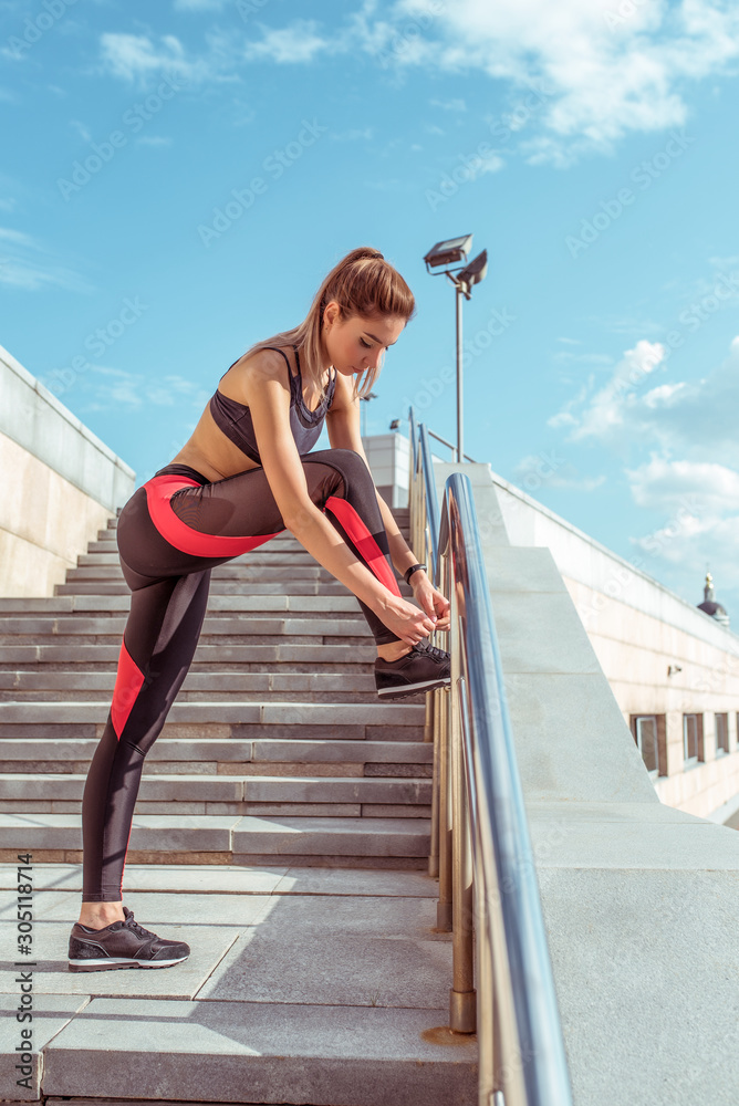 Beautiful athletic girl ties shoelaces on sneakers, jogging in the morning, stairs in the background in the summer, motivation lifestyle. Sportswear leggings top, tanned figure.