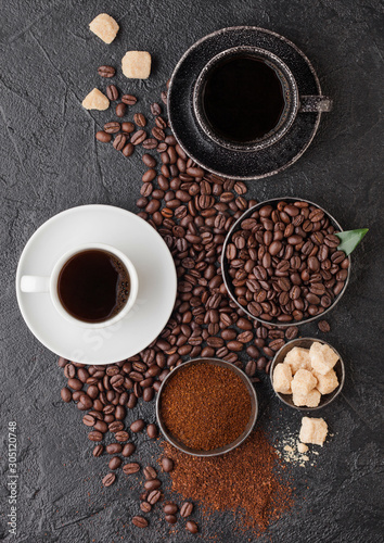 Black and white cups of fresh raw organic coffee with beans and ground powder with cane sugar cubes with coffee tree leaf on black background. Top view.