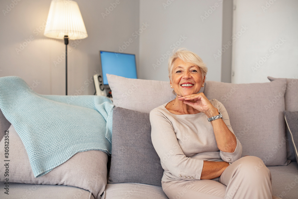 Indoor portrait of attractive happy 60 year old senior woman with pleasant smile relaxing on grey sofa in her modern apartment looking in anticipation while waiting for her children or grandchildren Indoor portrait of attractive happy 60 year old senior woman with pleasant smile relaxing on grey sofa in her modern apartment looking in anticipation while waiting for her children or grandchildren