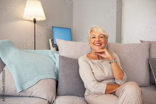 Indoor portrait of attractive happy 60 year old senior woman with pleasant smile relaxing on grey sofa in her modern apartment looking in anticipation while waiting for her children or grandchildren