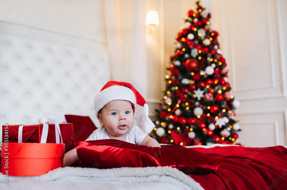 Baby in costume of Santa Claus with gift box lie on the bed in Christmas living room. Baby in costume of Santa Claus with gift box lie on the bed in Christmas living room.