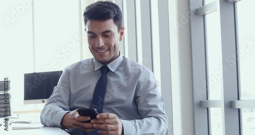 Handsome Caucasian businessman sitting and using smartphone for texting some message to his client in modern office.