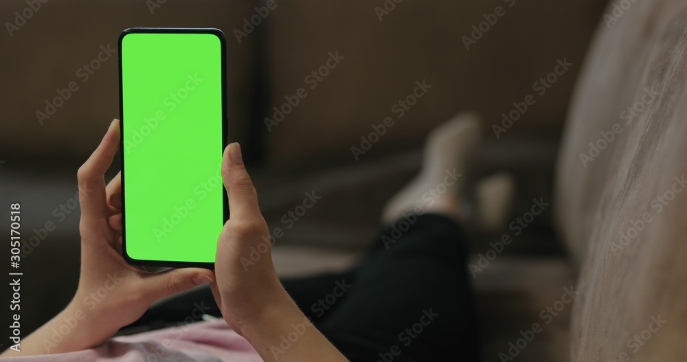 Young woman lying on a couch and using smartphone with vertical green screen