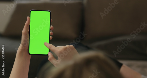 Young woman lying on a couch and using smartphone with vertical green screen