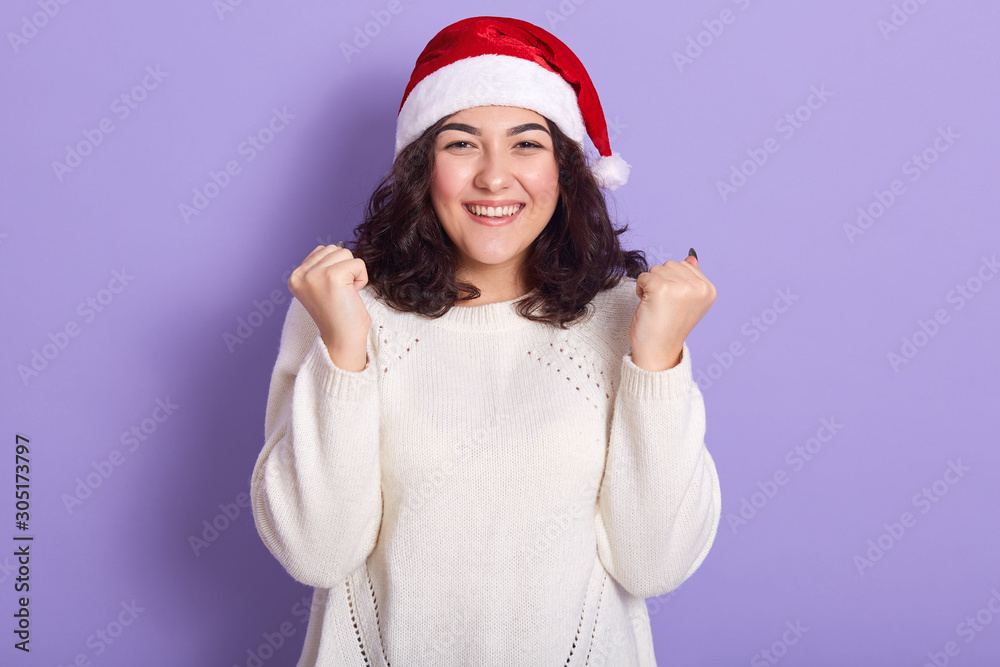 Beautiful female model wearing red santa claus and white sweater, raising her fists, smiling sincerely, having curly black hair, standing isolated over lilac background in studio, waiting for holiday. Beautiful female model wearing red santa claus and white sweater, raising her fists, smiling sincerely, having curly black hair, standing isolated over lilac background in studio, waiting for holiday.