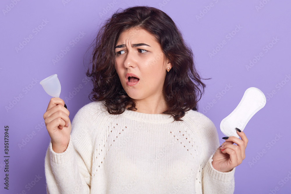 Studio shot of female holding menstrual cup and sanitarypad in hands over bright lilac background, does not know what to choose, stands with open mouth. Feminine hygiene product during period concept. Studio shot of female holding menstrual cup and sanitarypad in hands over bright lilac background, does not know what to choose, stands with open mouth. Feminine hygiene product during period concept.