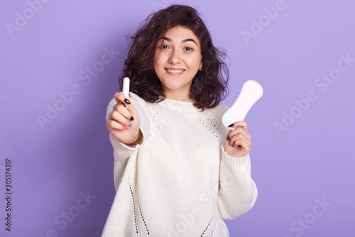 Portrait of cheerful attractive young lady holding tampon and sanitary napkin, making choices during menstruation, making preferance to tampon, looking directly at camera. Menstruation concept. photo