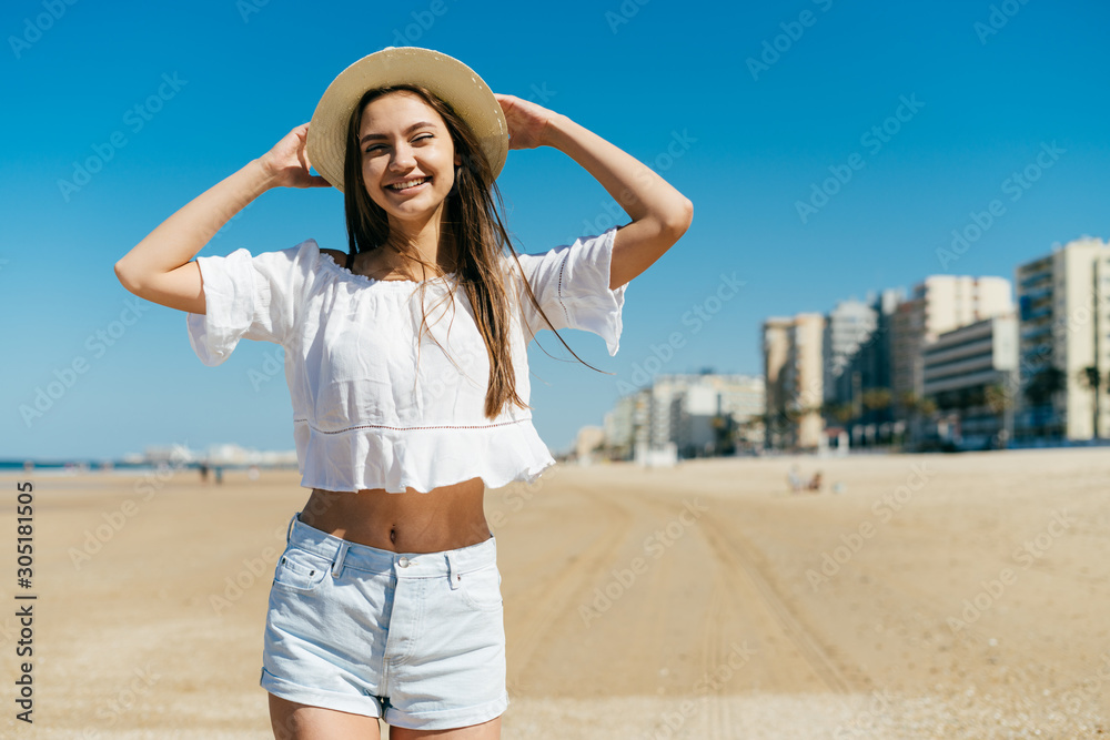 joyful girl smiles with complacency and holds her hat on the beach, away from the building joyful girl smiles with complacency and holds her hat on the beach, away from the building