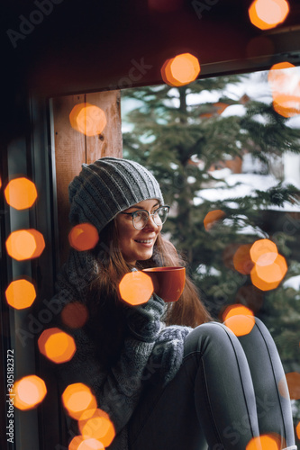 Beautiful woman holding and drinking a cup of coffee or cocoa in gloves sitting home by the window. Blurred winter snow tree background. Morning, coziness, winter and people concept