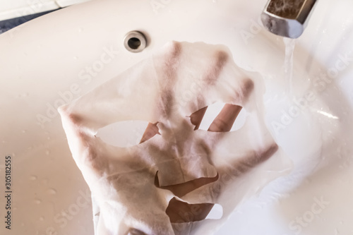 Woman hand holding a white sheet asian mask over a sink with the running water, skin care concept, washing sheet mask - Image