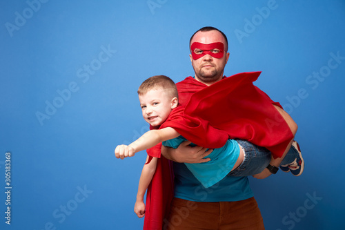 Man in mask and flying son superheroes in red cloaks on blank blue background