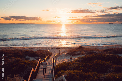 Young woman walking down the beach staircase and path to the sea with the orange sunset over the beautiful ocean in Perth, Western Australia. 