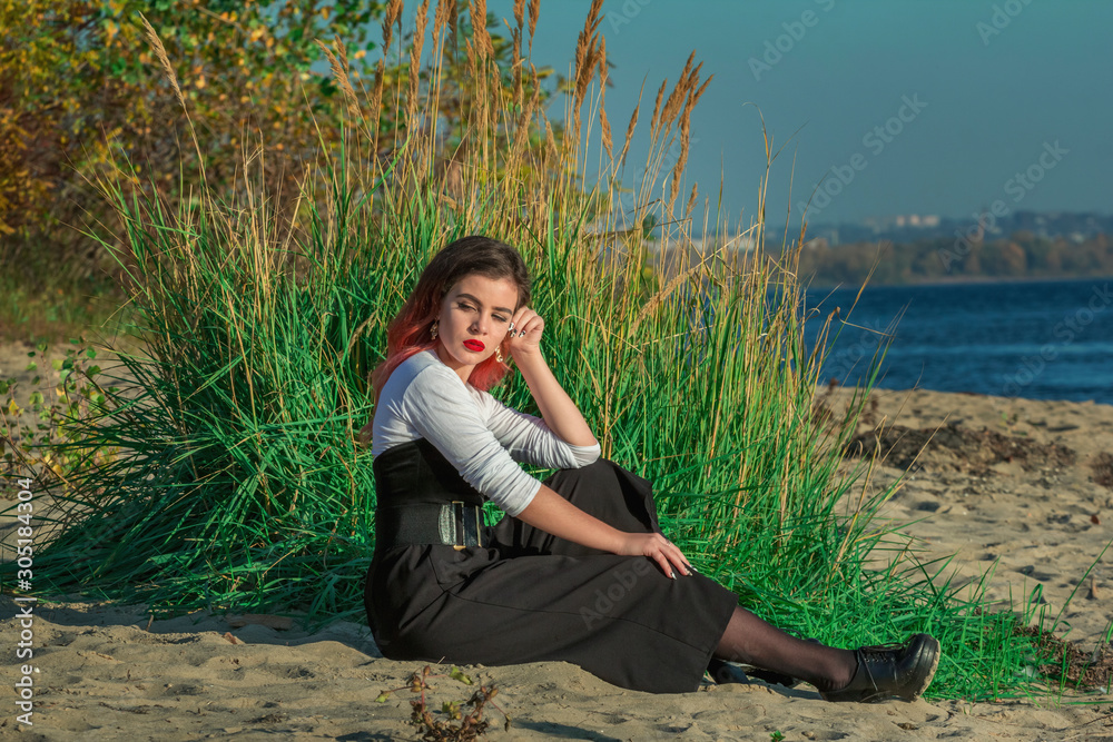 Portrait of beautiful stylish young woman with bright make-up, wearing white top and short fashionable culottes, sitting on a sand on river bank. 