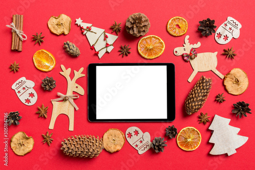 Top view of digital tablet, red background decorated with festive toys and Christmas symbols reindeers and New Year trees. Holiday concept