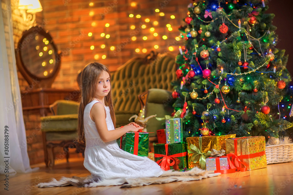 Cute little girl sits on the floor in the evening in the room near the festive Christmas tree, holds a gift box in her hands. Christmas and New Year.