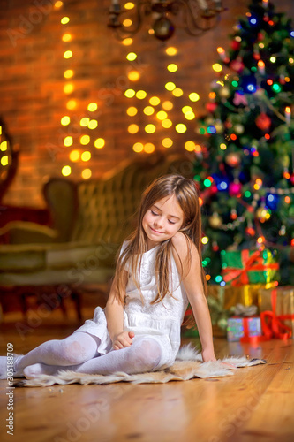 Cute little girl sits on the floor in the evening in the room near the festive Christmas tree and dreams. Christmas and New Year.