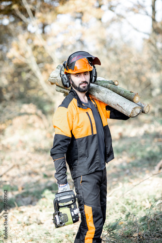 Portrait of a professional lumberman in protective workwear with a chainsaw and wooden logs during the work on logging in the pine forest