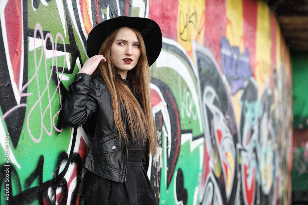 Obraz premium Young beautiful girl in a hat and with a dark make-up outside. Girl in the Gothic style on the street. A girl walks down the city street in a leather waistcoat with a phone.