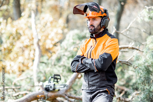 Waist-up portrait of a professional lumberman in harhat and protective workwear standing in the pine forest