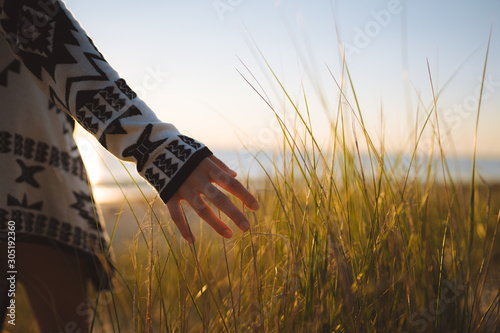 Fototapeta Naklejka Na Ścianę i Meble -  Young woman wearing a jersey with her hand exploring the sand dune grass on the beach with the sunset in the background over the ocean, Cottesloe, Perth, Western Australia.