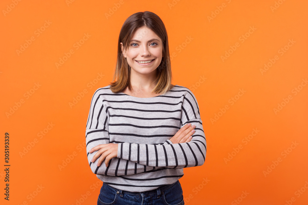 Portrait of cute happy young woman with brown hair in long sleeve shirt standing with crossed arms and looking at camera with kind toothy smile. indoor studio shot isolated on orange background Portrait of cute happy young woman with brown hair in long sleeve shirt standing with crossed arms and looking at camera with kind toothy smile. indoor studio shot isolated on orange background