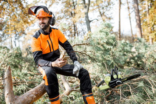 Portrait of a professional lumberman in protective workwear sitting with mobile phone on the felled tree, resting after the hard work in the forest