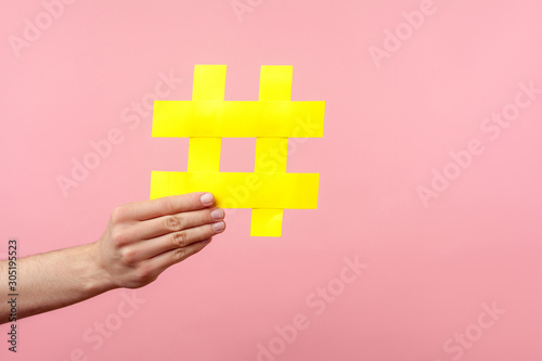 Closeup of male hand holding large yellow hashtag sign, concept of marketing and promotion in internet, followers in social media, trend content. indoor studio shot isolated on pink background