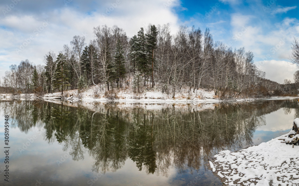 Winter landscape with forest on the lake, clouds in the sky and reflection