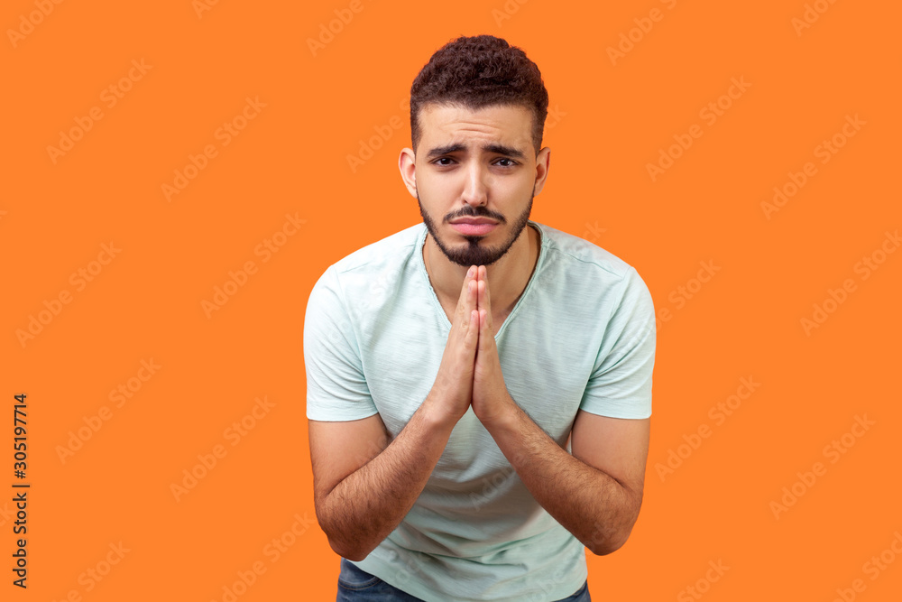 Please, I'm begging! Portrait of upset brunette man with beard in white t-shirt holding arms in prayer, asking help or forgiveness with imploring eyes. indoor studio shot isolated on orange background Please, I'm begging! Portrait of upset brunette man with beard in white t-shirt holding arms in prayer, asking help or forgiveness with imploring eyes. indoor studio shot isolated on orange background