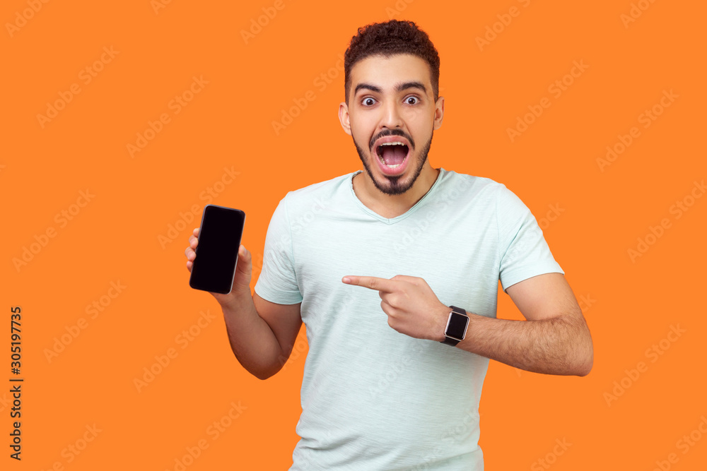 Portrait of shocked brunette man with beard in white t-shirt pointing at cellphone and looking at camera with amazement, surprised about mobile data tariffs. studio shot isolated on orange background Portrait of shocked brunette man with beard in white t-shirt pointing at cellphone and looking at camera with amazement, surprised about mobile data tariffs. studio shot isolated on orange background