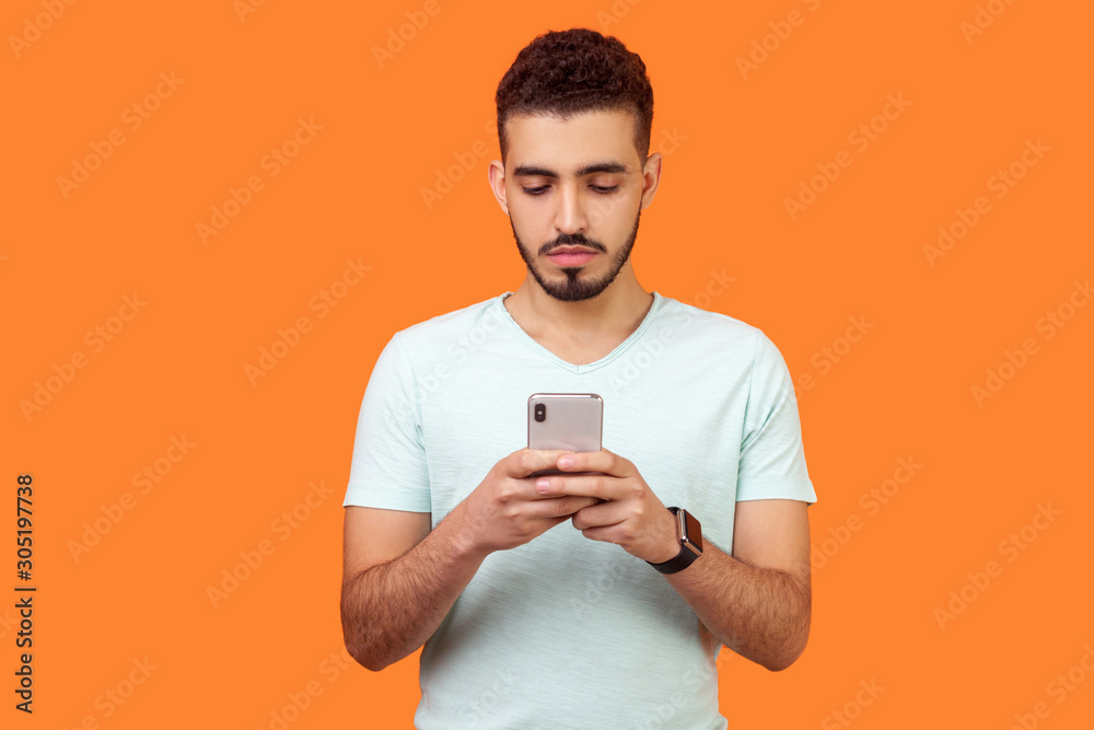 Portrait of serious attentive brunette man with beard in casual white t-shirt using cellphone, texting chatting in social media or surfing mobile web. indoor studio shot isolated on orange background Portrait of serious attentive brunette man with beard in casual white t-shirt using cellphone, texting chatting in social media or surfing mobile web. indoor studio shot isolated on orange background