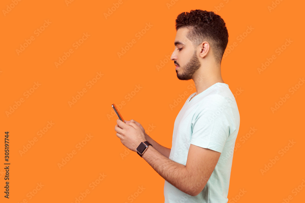 Side view of serious calm brunette man with beard in white t-shirt using cellphone, texting chatting in social media or surfing mobile web. empty copy space for advertisement, indoor studio shot