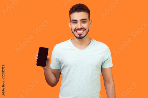 Portrait of positive brunette man with beard in white t-shirt holding cellphone and smiling, looking satisfied with device or mobile data tariffs. indoor studio shot isolated on orange background