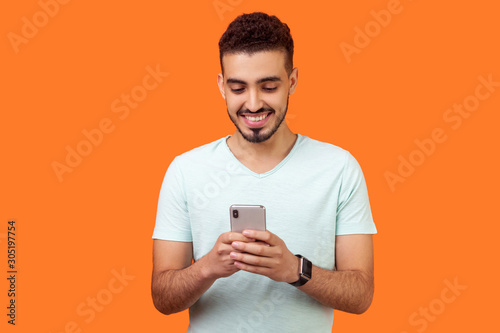 Portrait of cheerful brunette man with beard in white t-shirt using cellphone and smiling, reading good news message, enjoying mobile application. indoor studio shot isolated on orange background © khosrork