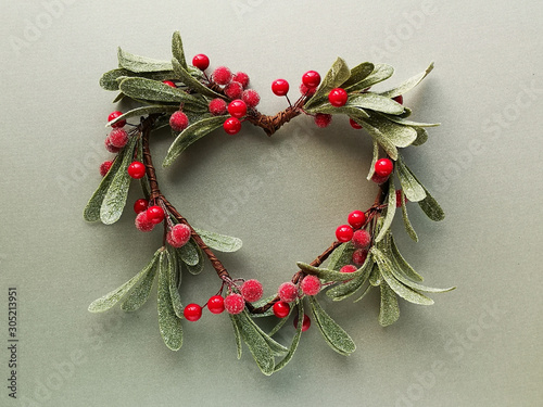 Decorative mistletoe wreath with frosted leaves and red berries shaped as a heart. Flat lay, top view on metallic green paper.