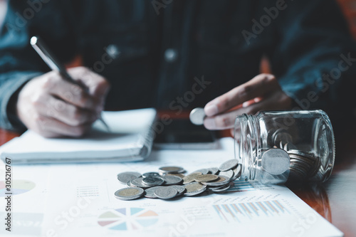 Man is taking notes of saving money in a notebook, with a money jar and a business report on the table. © zeeclock1