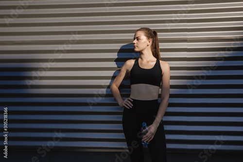 Healthy female standing outdoors and drinking water after exercising session. Fitness woman taking a break after workout