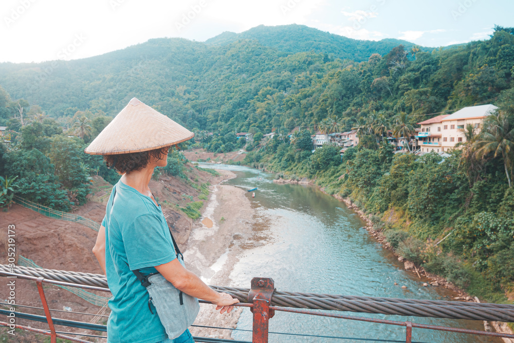 Woman with traditional hat looking at view on obsolete footbridge over river in Muang Khua village in North Laos, travel destination in South East Asia. Toned teal orange style