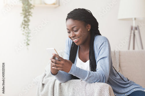 Happy black woman using smartphone at home, relaxing on sofa