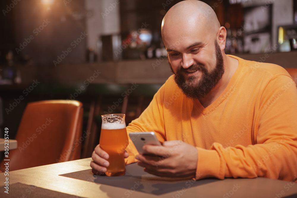 Cheerful bearded man using his smart phone while drinking beer at the pub Cheerful bearded man using his smart phone while drinking beer at the pub