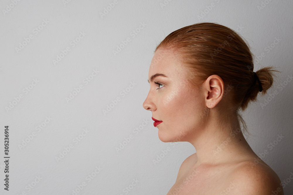 Side portrait of pretty young woman with naked shoulders and red lips looking at the camera and smiling, posing on the gray wall. Isolated. Side portrait of pretty young woman with naked shoulders and red lips looking at the camera and smiling, posing on the gray wall. Isolated.
