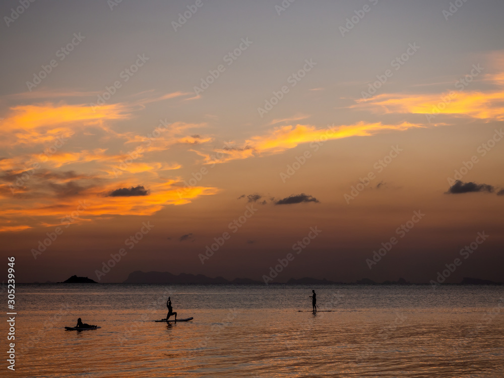 Silhouettes of people on sup boards in the rays of the setting sun against a background of clouds. Koh Phangan. Thailand.