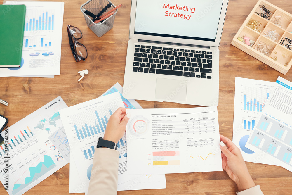 Obraz premium Above view background of unrecognizable businesswoman holding statistics data graph while working at wooden desk with documents laid out and words Marketing Strategy on laptop screen, copy space