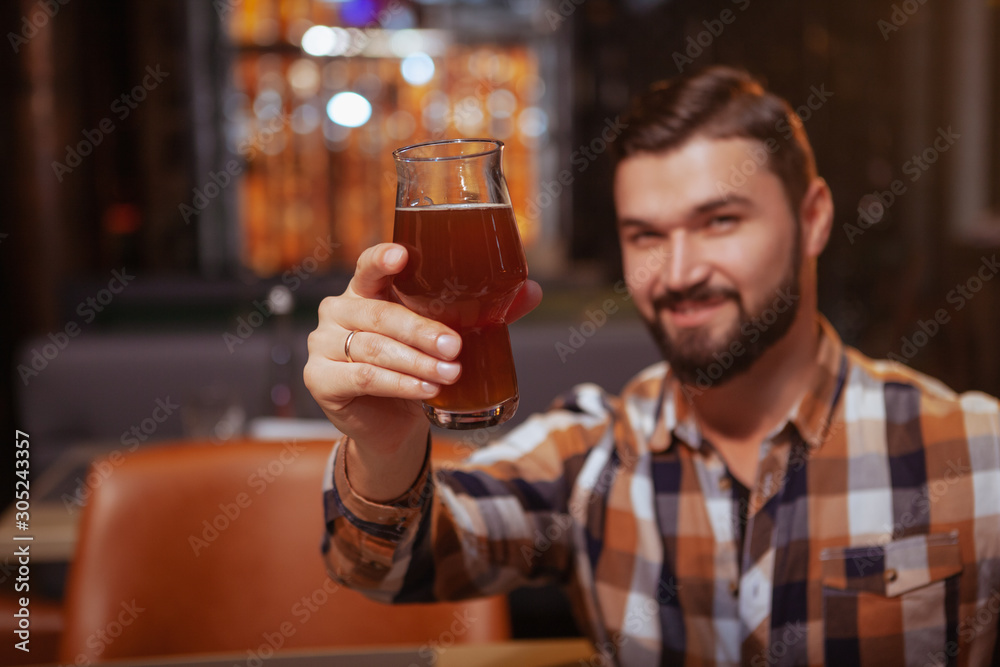 Selective focus on a beer glass in the hand of happy handsome bearded man. Cheerful man toasting with his glass to the camera