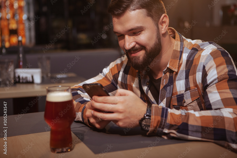Happy handsome man texting on his smart phone at beer pub. Attractive cheerful man using his phone while drinking beer at the bar Happy handsome man texting on his smart phone at beer pub. Attractive cheerful man using his phone while drinking beer at the bar