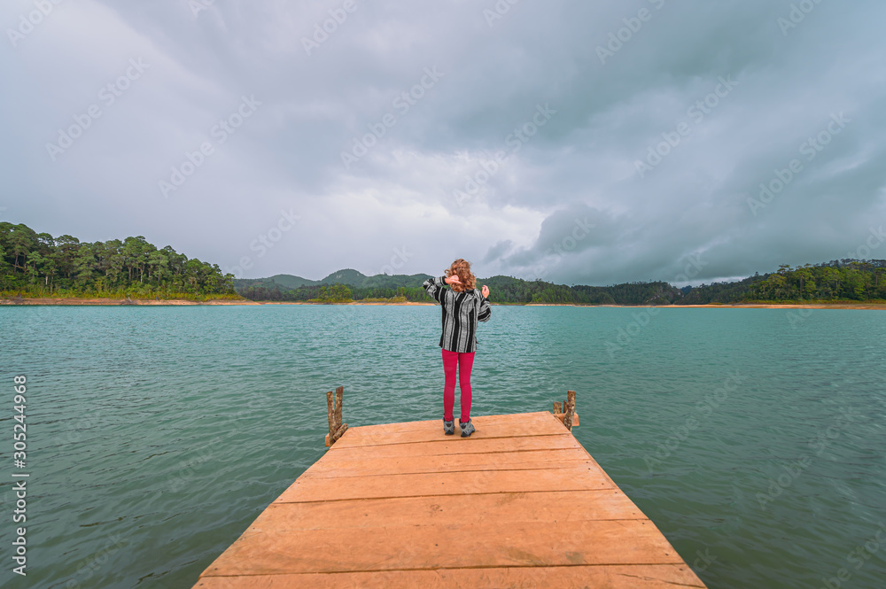 Young blonde woman on a pier watching a turquoise lake and background mountains with forest