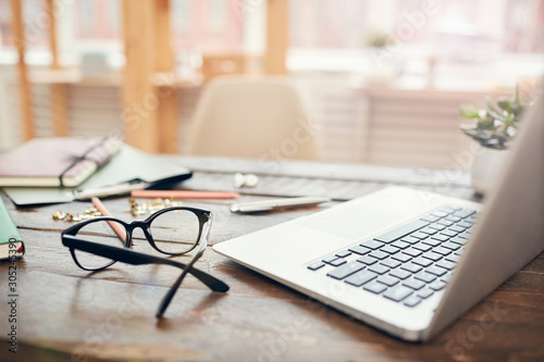 Background image of business office with laptop and supplies on wooden desk, focus on black hard rim glasses in foreground, copy space