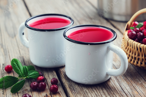 Two enameled mugs of or cranberry juice or mors. A cranberry drinks, basket of berries and pan on background.